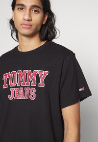 Juodas medvilninis marškinėlis su ryškiu raudonu ir baltu užrašu "TOMMY JEANS". Turi trumpas rankoves ir mažą logotipą kairėje rankovėje.