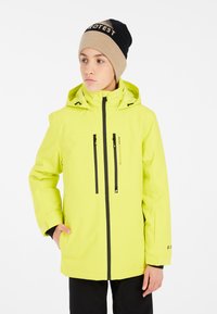 Veste imperméable jaune vif avec capuche, dotée de poches zippées et d'accents noirs. Portée avec un bonnet beige et noir.