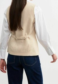 Beige linneväst med en insnörd detaljer bak, buren över en vit skjorta, i kombination med mörkblå jeans. Har en mjuk textur och skräddarsydd design.