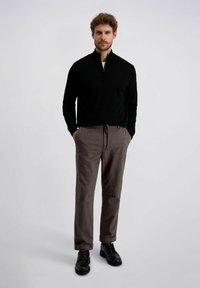 Pull noir à demi-zip avec manches longues, associé à un pantalon à motifs marron et des chaussures à lacets noires. Le modèle se tient devant un fond neutre.