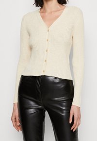 Cárdigan crema acanalado con escote en V, que cuenta con un cierre de botones en la parte delantera y mangas largas. Combinado con pantalones negros brillantes.