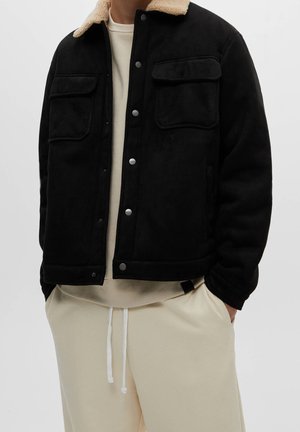 Muž v černé zapínací bundě s béžovým límcem z beránčí kožešiny přes béžovou mikinu a béžové kalhoty se stahovací šňůrkou. Ruce v kapse.