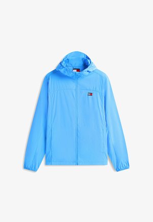 Coupe-vent à capuche bleu clair avec fermeture éclair frontale et petit logo rectangulaire rouge, blanc et bleu sur la poitrine gauche.