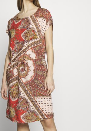 Frau trägt ein kurzärmliges Kleid mit einem rot-beigen Paisley- und Blumenmuster, geraffter Taille mit einem Bindegürtel, vor weißem Hintergrund.