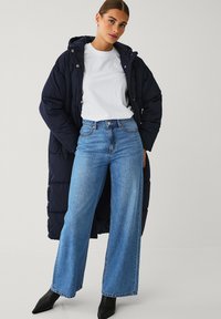 Marine gewatteerde lange jas, witte t-shirt met ronde hals, blauwe wijde jeans en zwarte haklaarzen. Opmerkelijke details zijn knopen en naden.