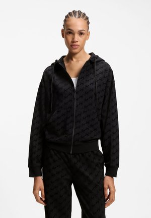 Donna con capelli intrecciati che indossa una felpa con zip nera e pantaloni abbinati con un motivo sottile e ripetuto del logo, in piedi su sfondo bianco.