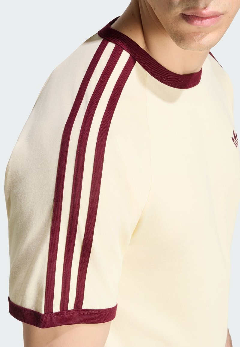 Hombre con camiseta color crema con cuello burdeos, puños de las mangas y tres franjas burdeos a lo largo del hombro y la manga.