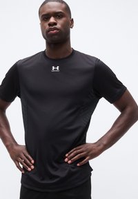 Αθλητικός άντρας στέκεται με τα χέρια στα ισχία, φοράει μαύρο κοντομάνικο μπλουζάκι Under Armour, σε απλό λευκό φόντο.