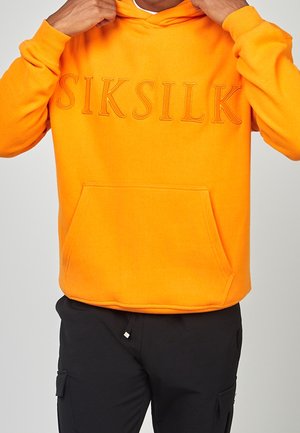 Osoba poprawiająca jasnopomarańczową bluzę z kapturem z przednią kieszenią i podniesionym logo "SIKSILK", ubrana w czarne spodnie z boczną kieszenią.