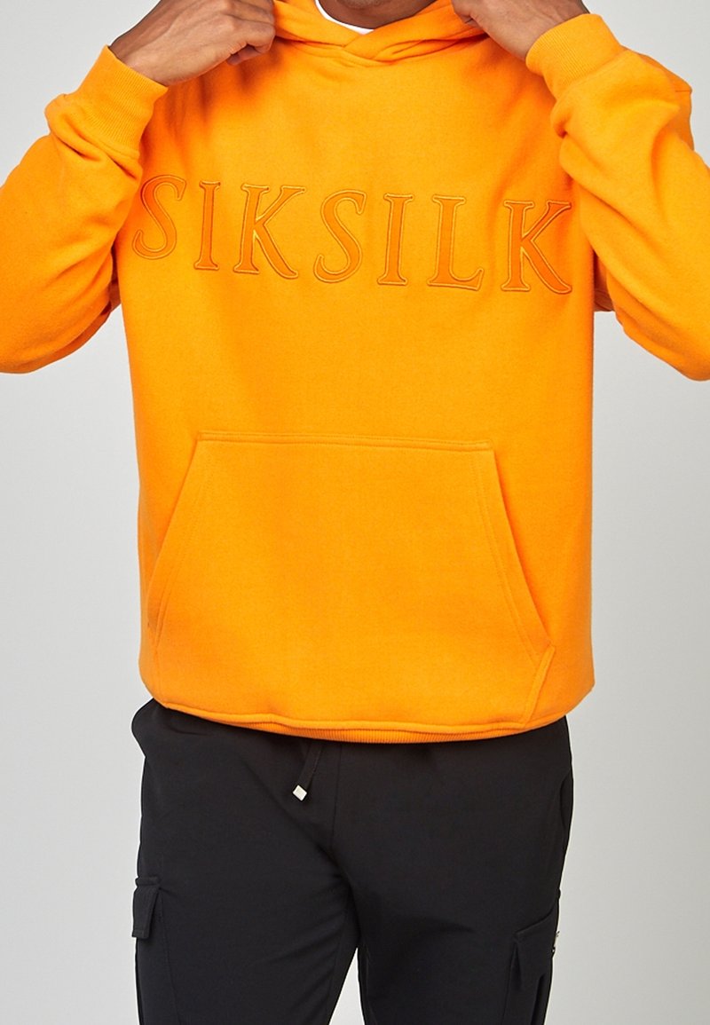 Personne ajustant un sweat à capuche orange vif avec poche devant et logo "SIKSILK" en relief, portant un pantalon noir avec une poche latérale.