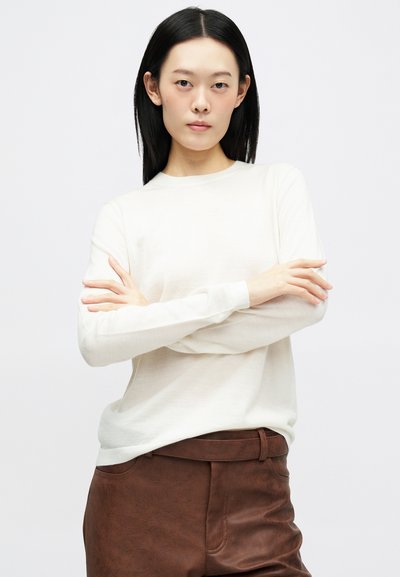 Jovem mulher com cabelo preto liso, vestindo uma blusa branca de mangas longas e calças de couro castanhas, de pé com os braços cruzados.