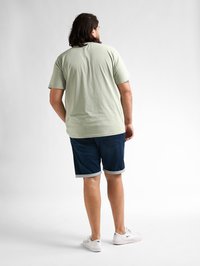 Lichtgroen t-shirt met korte mouwen, donkere spijker shorts met opgerolde zoom, witte sneakers. Van achteren weergegeven tegen een neutrale achtergrond.