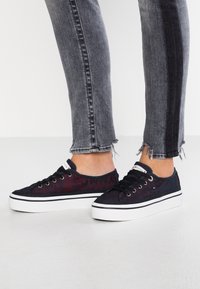 Leinenschuhe mit einem gewebten navyblauen Obermaterial, roten Logo-Details, schwarzen Schnürsenkeln und einer weißen Plateausohle. Kombiniert mit abgetragenen grauen Jeans.
