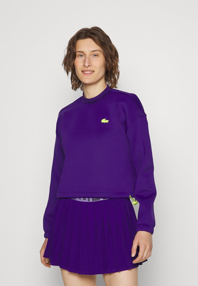 Personne portant un haut à manches longues violet avec un logo jaune et une jupe plissée assortie, se tenant devant un fond clair et uni.