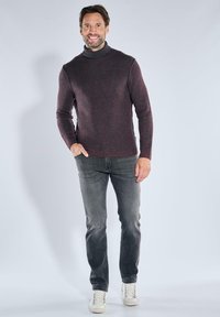 Ein lächelnder Mann steht mit einer Hand in der Tasche, trägt einen dunkel gerippten Rollkragenpullover, graue Jeans und weiße Turnschuhe vor einem schlichten Hintergrund.