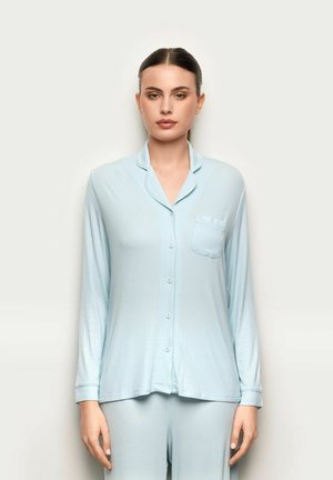 Mujer con blusa de botones de manga larga color azul pálido y pantalones a juego, de pie contra un fondo claro y liso, mirando hacia adelante con expresión neutral.