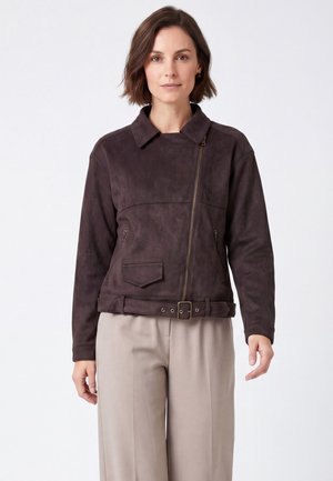 Frau trägt eine dunkelbraune Wildlederjacke mit asymmetrischem Reißverschluss und Gürtel, kombiniert mit hellbeigen Hosen, stehend vor weißem Hintergrund.