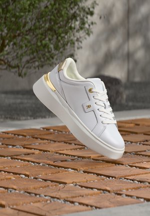 Zapatilla blanca con detalles dorados flota sobre baldosas decoradas al aire libre, con vegetación y una pared de hormigón desenfocada al fondo.