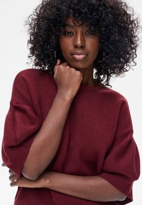 Bordowy, luźny sweter z krótkimi, szerokimi rękawami; materiał o fakturze. Modelka ma kręcone włosy i opiera podbródek na dłoni, widoczne kolczyki.