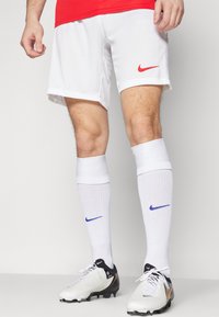 Weiße Fußballshorts von Nike mit rotem Logo, kombiniert mit weißen Socken mit blauen Logos und schwarz-weißen Fußballschuhen.