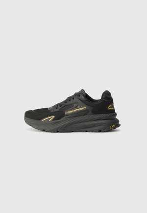 Baskets noires EA7 Emporio Armani avec semelle épaisse, dessus en mesh et cuir, détails logo dorés et lacets sur fond gris clair uni.