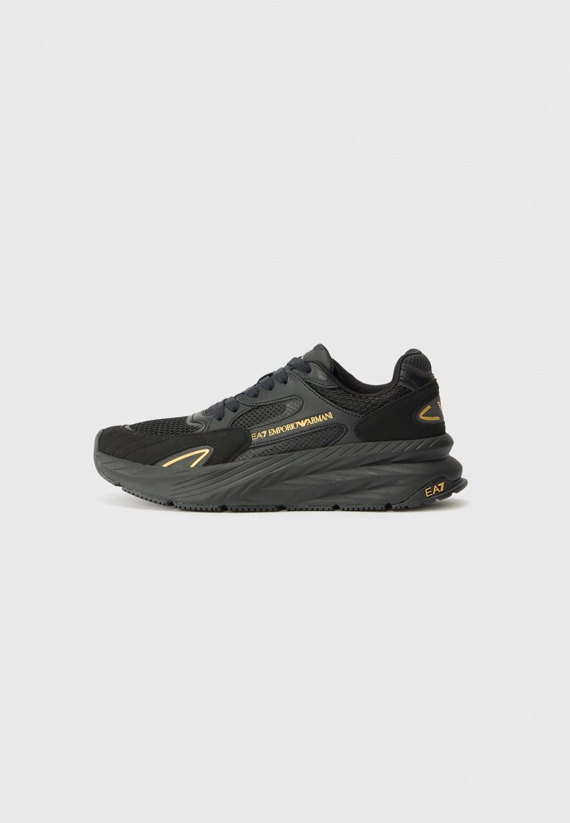 Baskets noires EA7 Emporio Armani avec semelle épaisse, dessus en mesh et cuir, détails logo dorés et lacets sur fond gris clair uni.
