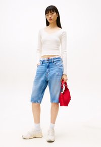 Lett hvit langermet topp, blå denimshorts, hvite joggesko, rød håndveske. Uformell antrekk med synlige merkevarer på midjebåndet.