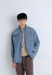 Veste en denim bleu clair avec deux poches poitrine, fermetures à bouton et coupe décontractée. Portée sur un haut beige, associée à un pantalon marron.