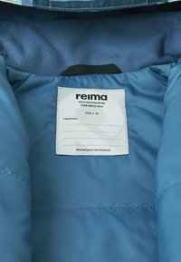 Interno di una giacca imbottita blu con tessuto liscio. Un'etichetta di cura mostra il marchio "Reima", la taglia "104 / 4T" e un messaggio per passarlo ad altri.