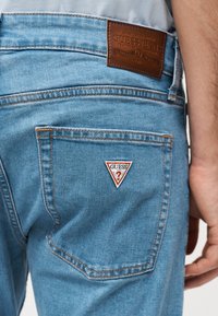 Φωτεινό μπλε τζιν με δερμάτινη ετικέτα GUESS Jeans στη μέση και τριγωνικό λογότυπο GUESS στην πίσω τσέπη.