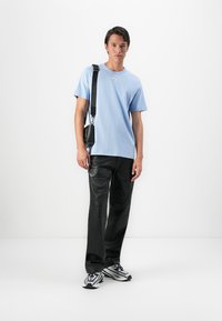 Ungdom som har på seg en lyseblå t-skjorte, svarte cargobukser, sølvfargede joggesko og en svart crossbody-veske, stående mot en hvit bakgrunn.