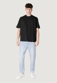 Camicia nera a maniche corte con texture e motivo a rombi, abbinata a jeans azzurri e sneakers bianche. Il modello è in piedi contro uno sfondo neutro.