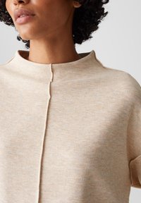 Krullend haar persoon draagt een lichtbeige geribbelde trui met een verticale naaddetail in het midden en een geribbelde mock neck.