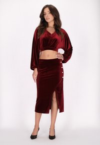 Fluwelen twee-delige set in dieprood. Bevat een cropped top met een V-hals en lange mouwen, gecombineerd met een rok met een zijslit en knopen.