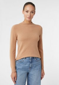 Hellbraun gestrickter Pullover mit hohem Kragen und langen Ärmeln, aus weichem Material. Kombiniert mit blauen, lockeren Jeans. Schlichtes, sauberes Design.