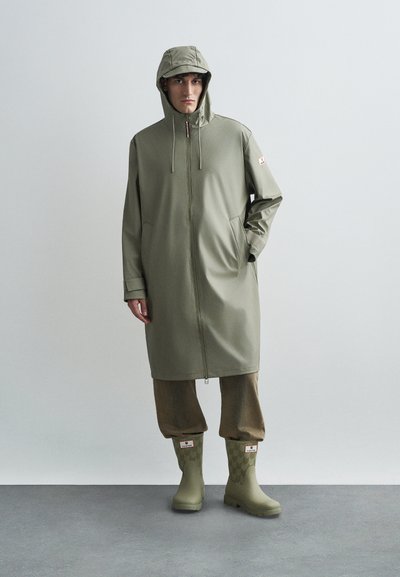 Moški v olivno zeleni dežni jakni s kapuco, svetlih hlačah in ustreznih olivno zelenih gumijastih škornjih, ki stoji pred enobarvno svetlo sivo ozadjem.