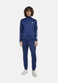Chándal navy con franjas laterales blancas, chaqueta con cremallera y pantalones a juego. Tejido suave, con el logo de Adidas en el pecho y en el muslo.