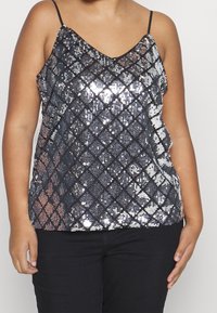 Top con lentejuelas negras brillantes y un patrón de diamantes, escote en V y tirantes delgados ajustables. Llevado sobre pantalones negros, la superficie texturizada refleja la luz.
