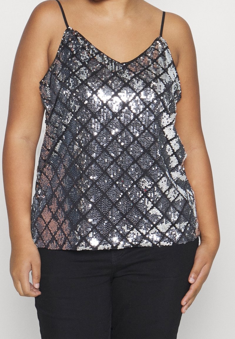 Top con lentejuelas negras brillantes y un patrón de diamantes, escote en V y tirantes delgados ajustables. Llevado sobre pantalones negros, la superficie texturizada refleja la luz.