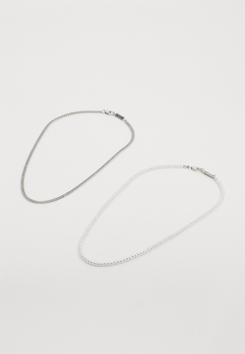 Dos collares: uno es una cadena de plata con un diseño de eslabones planos, y el otro es un collar de cuentas blancas con una textura suave.