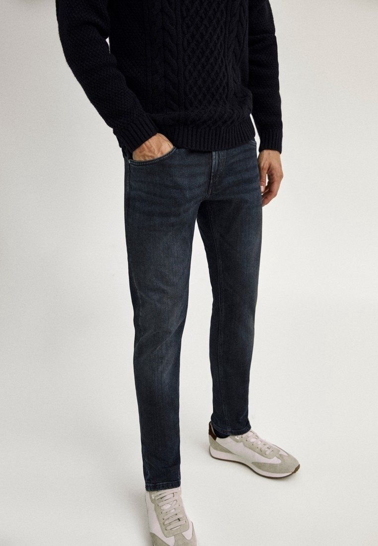 Homme portant un jean slim foncé, un pull en maille torsadée noir et des baskets blanches avec des touches beiges, debout avec une main dans la poche.