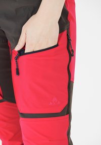 Rode en zwarte broek met een gestructureerde afwerking, voorzien van een ritszak aan de zijkant en een klein logo op het bovenbeen.