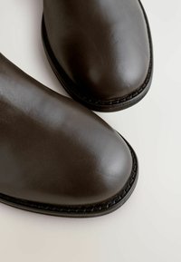 Stivali alla caviglia in pelle marrone scuro con una finitura liscia, punta arrotondata e cuciture nere a contrasto lungo il bordo della suola.