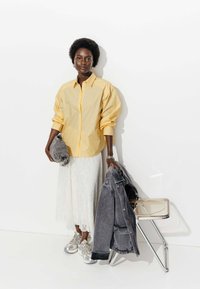 Chemise oversize à rayures jaunes, jupe en dentelle blanche, veste en denim grise et baskets argentées, avec une chaise transparente en arrière-plan.