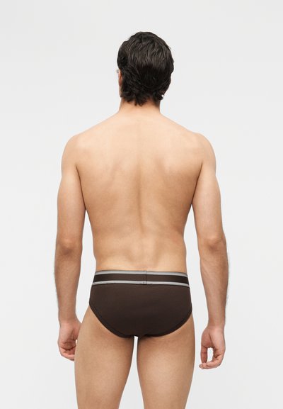 Homme aux cheveux foncés vu de dos portant des slips marron foncé avec une ceinture grise rayée sur un fond clair uni.