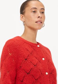 Rode gebreide cardigan met een diamanten patroon, ronde halslijn en witte knopen. De textuur lijkt zacht en delicaat.