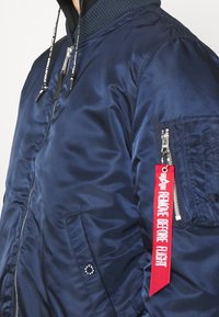 Veste bomber bleu marine avec poche zippée sur la manche, dotée d'une étiquette rouge indiquant "Remove Before Flight" et fermeture zippée à l'avant.