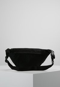 Banane en tissu noir de forme triangulaire, avec une sangle réglable et une poche zippée à l'avant. Texture mate et design minimaliste.