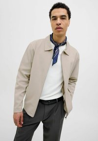 Lichtbeige zip-up jack met een kraag, gecombineerd met een witte trui en donkere broeken. Bevat een blauwe sjaal met patroon om de nek.