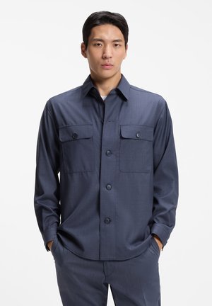 Giovane uomo che indossa una camicia blu scuro con bottoni e due tasche sul petto, in piedi con le mani nelle tasche contro uno sfondo semplice.